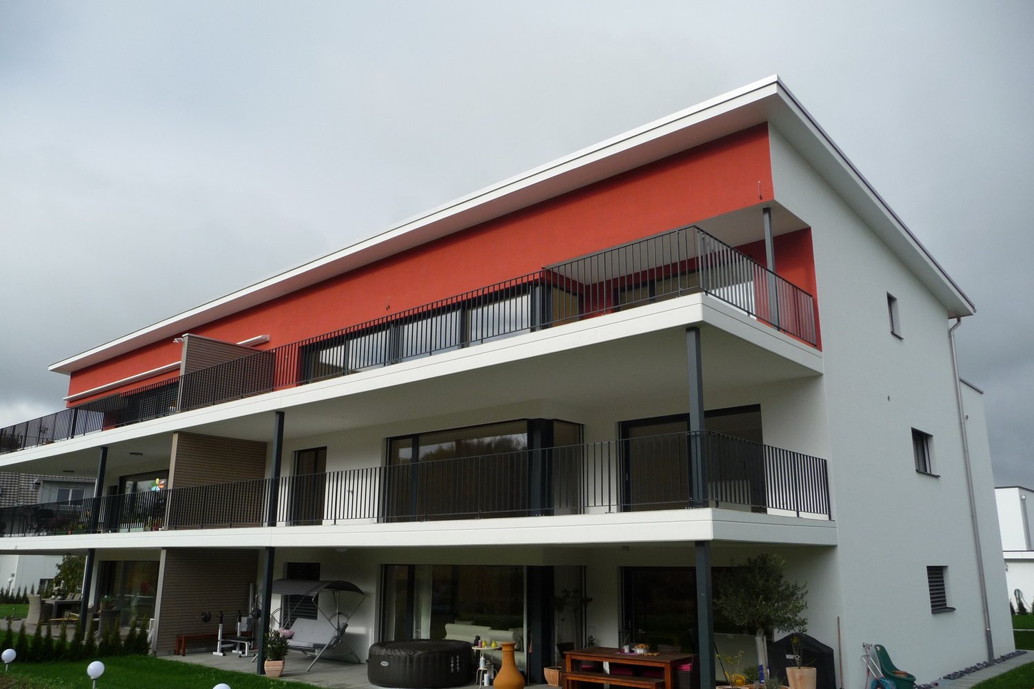 Modern 3-storey building, red and white exterior, large windows, black railings, balconies on each floor, garden with plants and outdoor furniture.