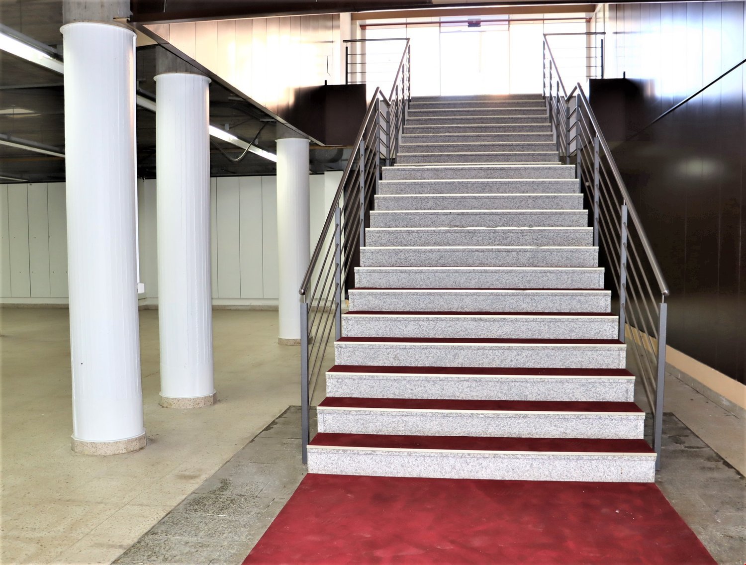 The image shows a set of stairs with a red carpet leading up to the second floor. The stairs are made of granite or a similar stone material, with metal railings on both sides. The stairs are located in a large open space with white columns supporting the