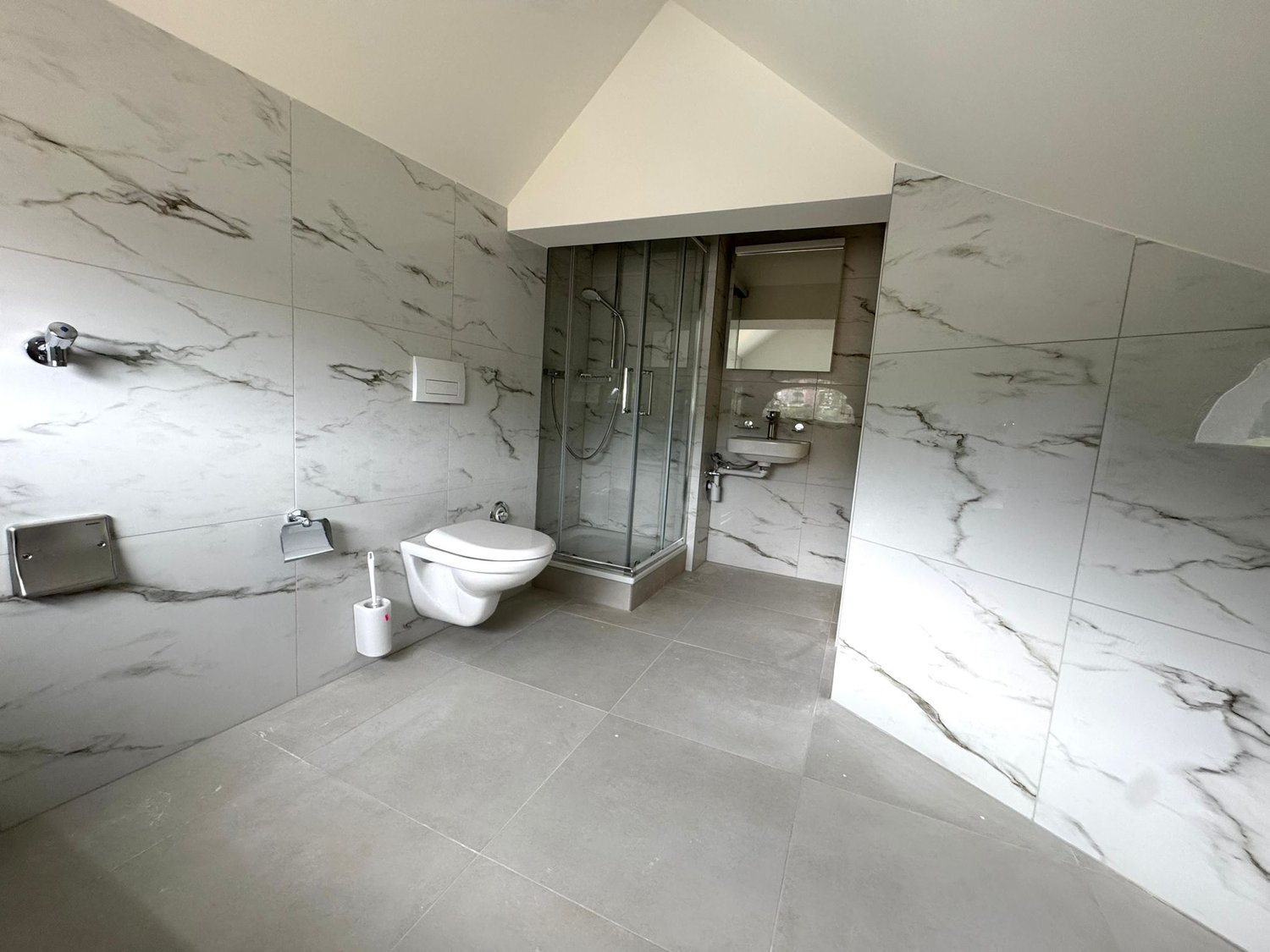 Spacious bathroom with marble-like wall tiles, glass shower enclosure, toilet, and vanity sink. The room has a modern and minimalist design.