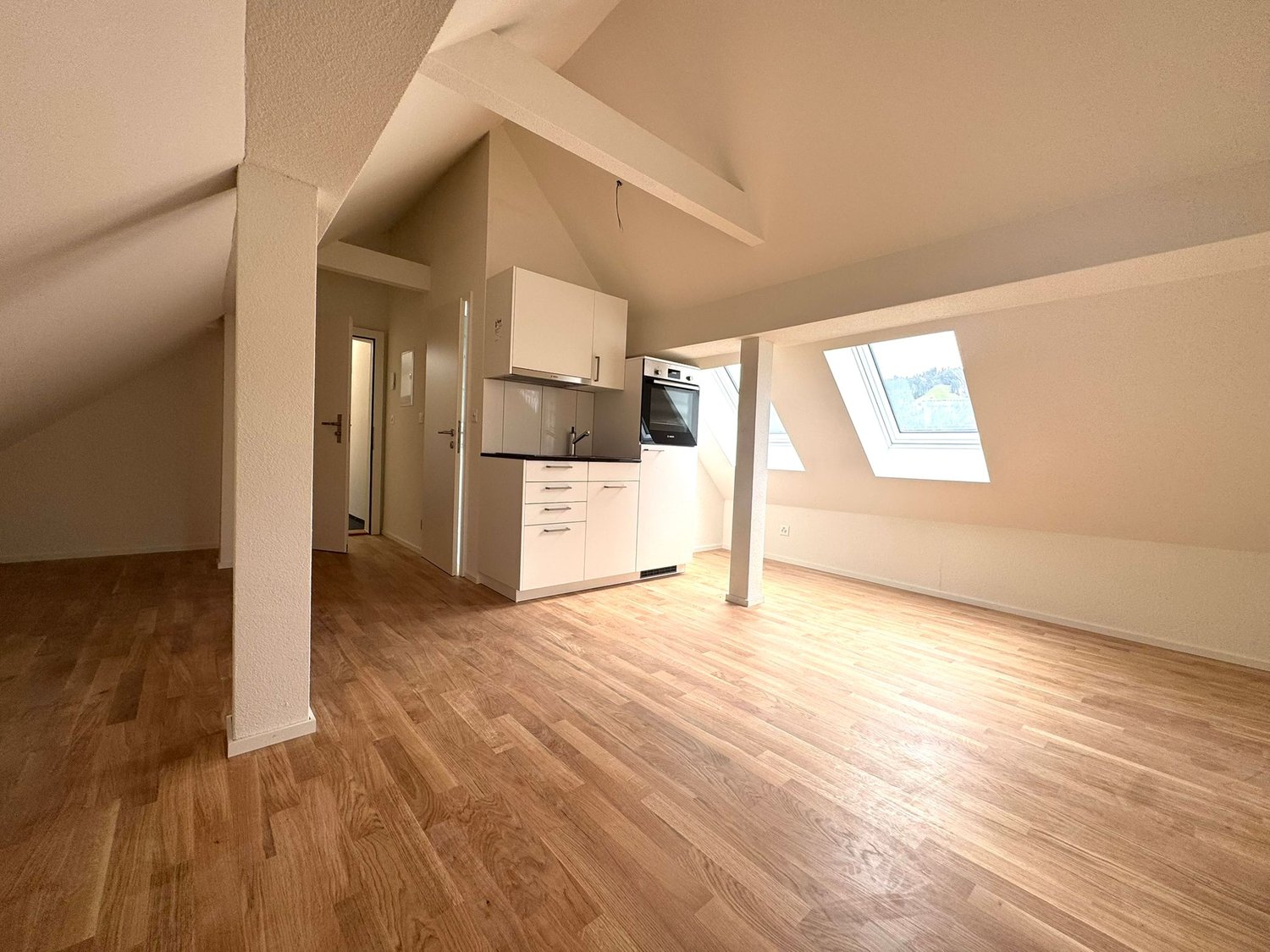 The image shows a studio apartment with an open floor plan. The space features hardwood flooring, white walls, and a slanted ceiling with exposed beams. The kitchen area is visible, with white cabinets and appliances. There are large windows that provide