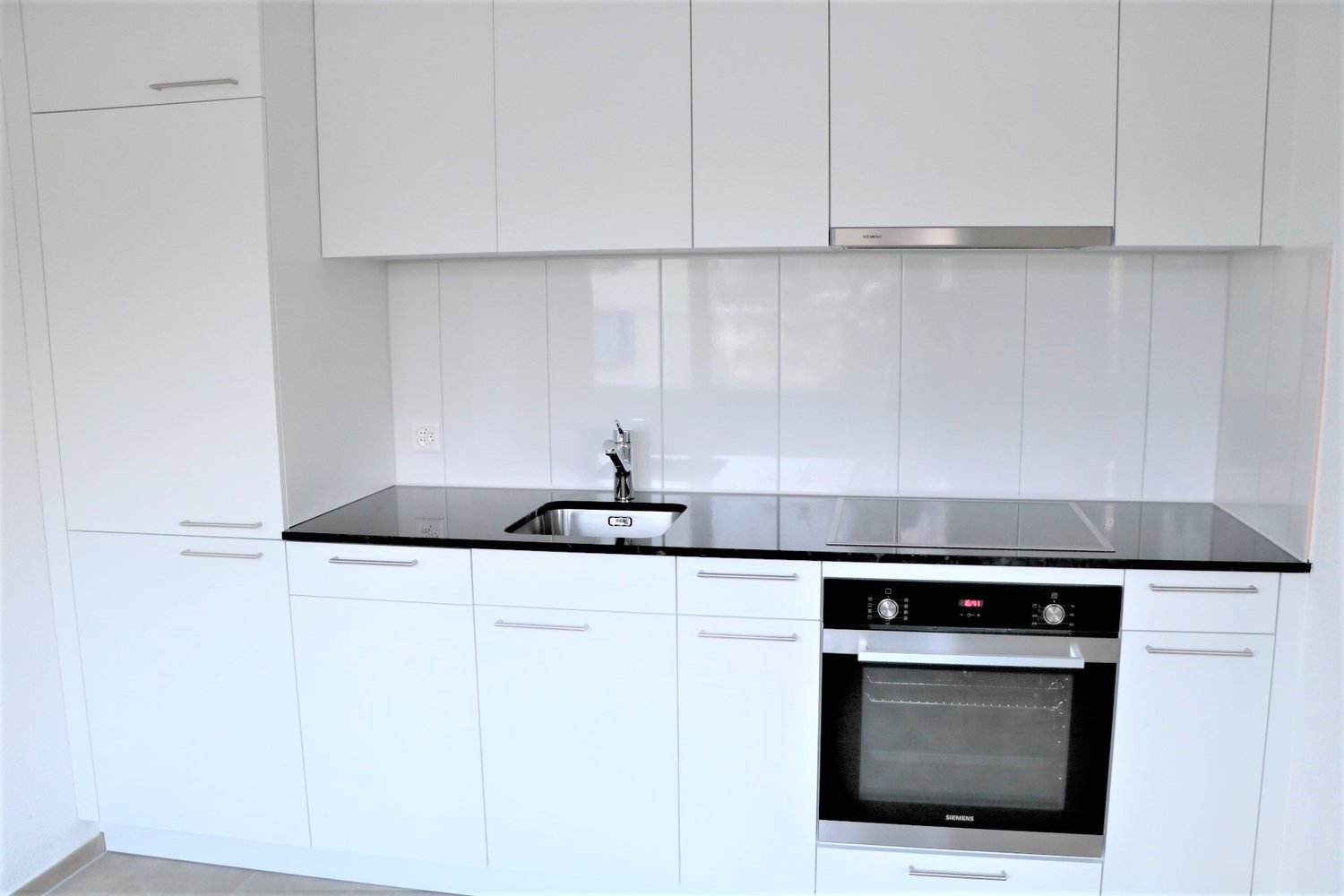 Modern, minimalist kitchen with white cabinets, black countertop, stainless steel sink, and built-in oven and dishwasher