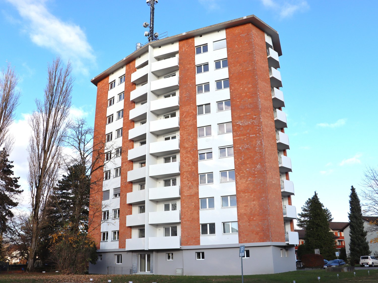 6-story building, brick facade, white balconies, telecommunication tower on top, small garden area