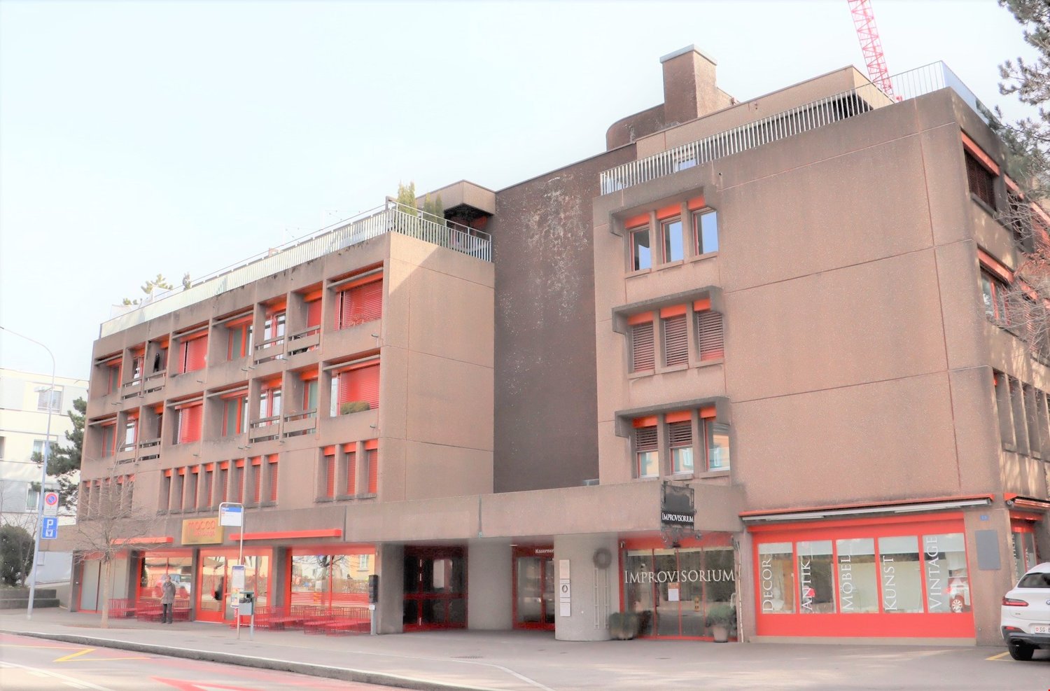 Multi-story commercial building with red accents, balconies, and retail spaces on the ground floor