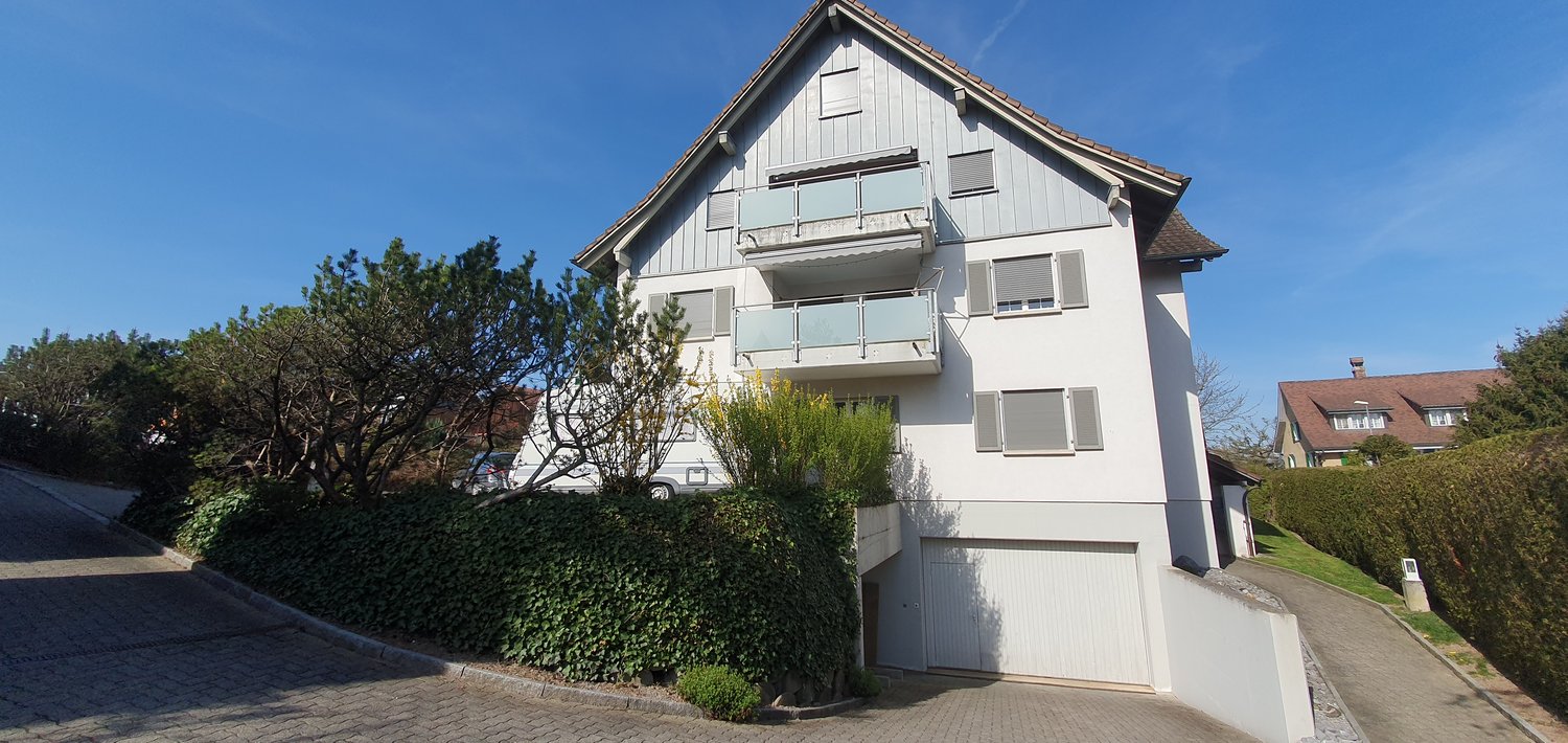 Two story house, white exterior, balcony, garage, driveway, paved sidewalk, hedges, trees