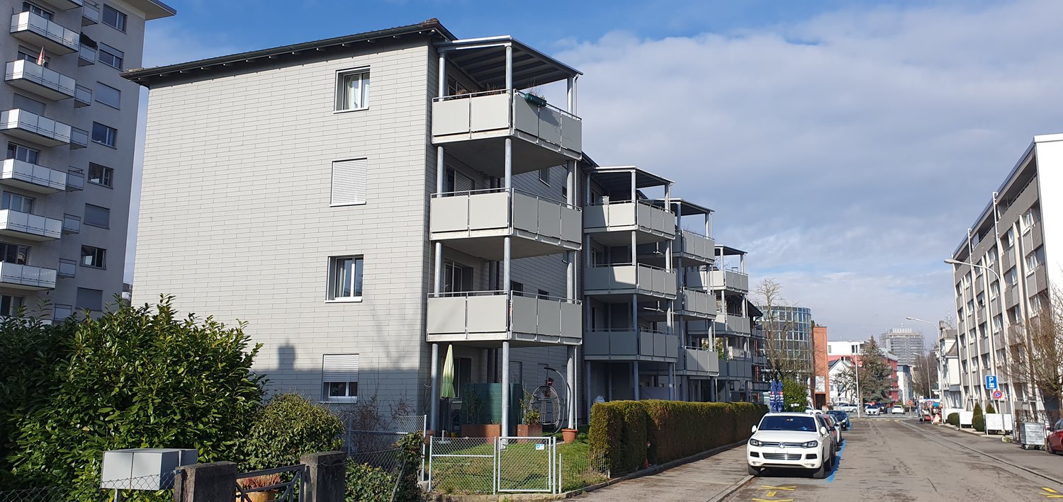 Multi-story apartment building, balconies on multiple floors, white brick facade, glass windows, hedges and greenery in front