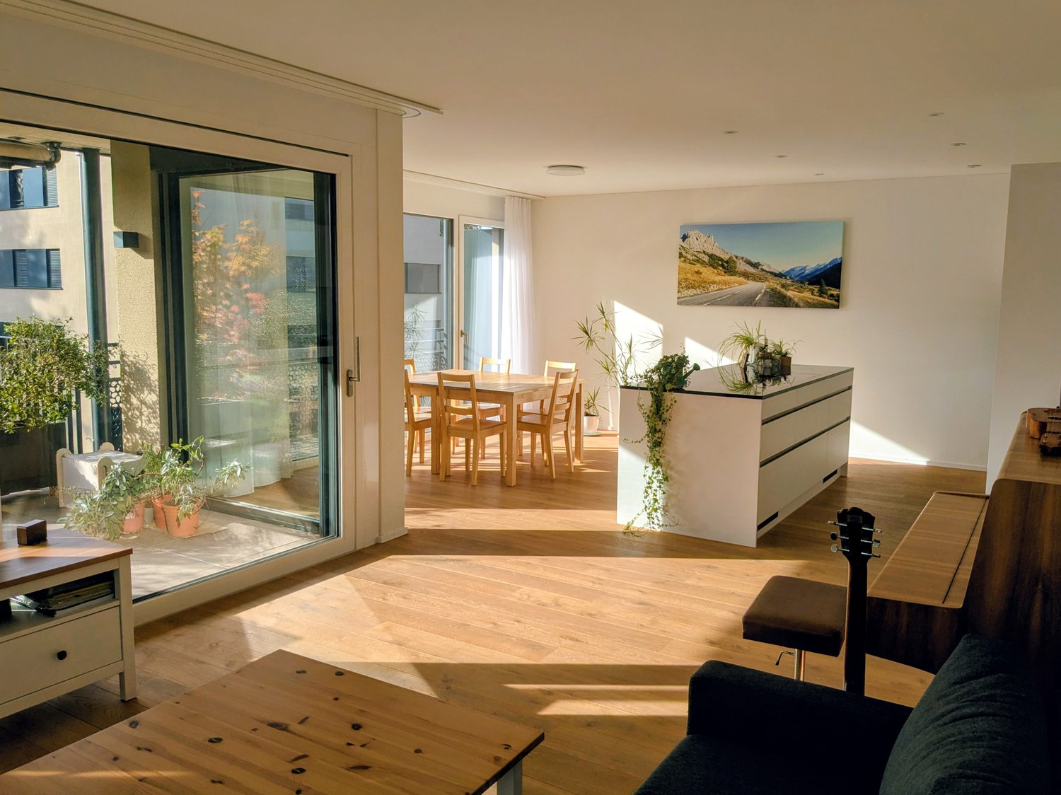 Wooden floor, white walls, large glass door leading to terrace, dining table with chairs, white kitchen counter, guitar, wooden stools, coffee table, potted plants, mounted painting
