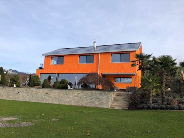 Two story house, orange paint, grey solar panels, large windows, stone wall with plants, green lawn, palm trees