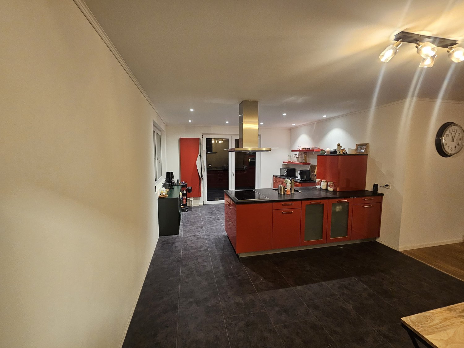 Modern kitchen with red cabinetry, black countertops, and a stove hood, equipped with a microwave, refrigerator, and various kitchenware. Large windows and wooden floor
