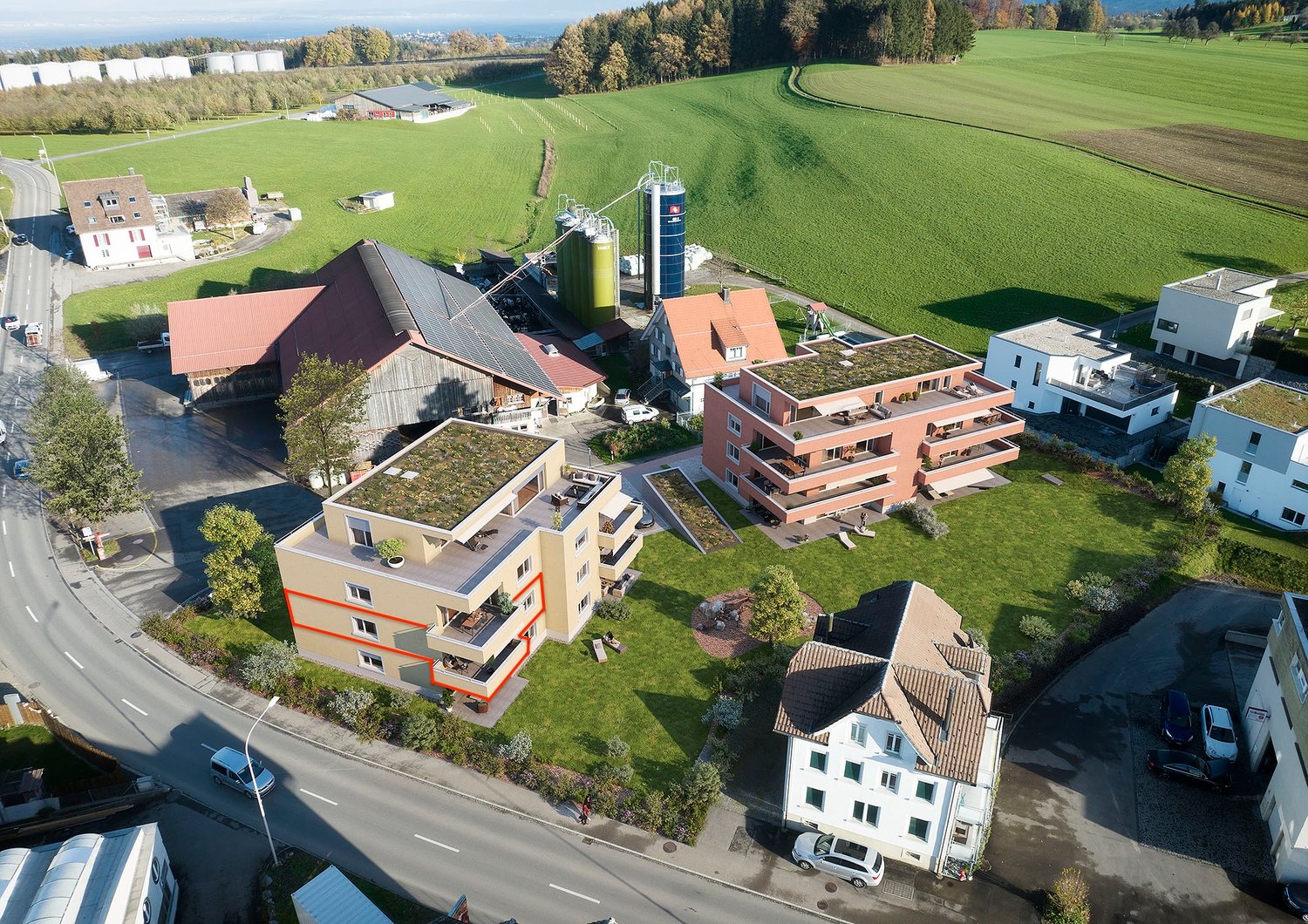 A complex of residential buildings, with various sizes and shapes, are located on a large green field with several trees and plants. Several cars are parked in the area, and some of them are parked in the driveway. The buildings have a mix of architectura