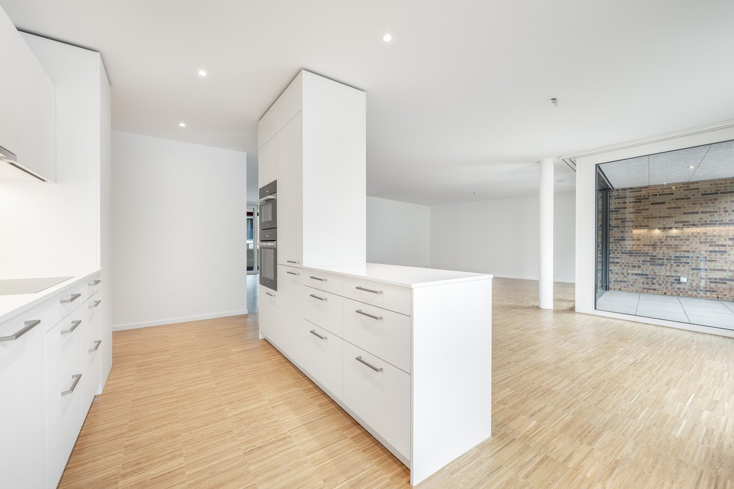 Minimalist modern kitchen with white cabinets, wooden floors, integrated oven, and sliding glass doors leading to a patio.