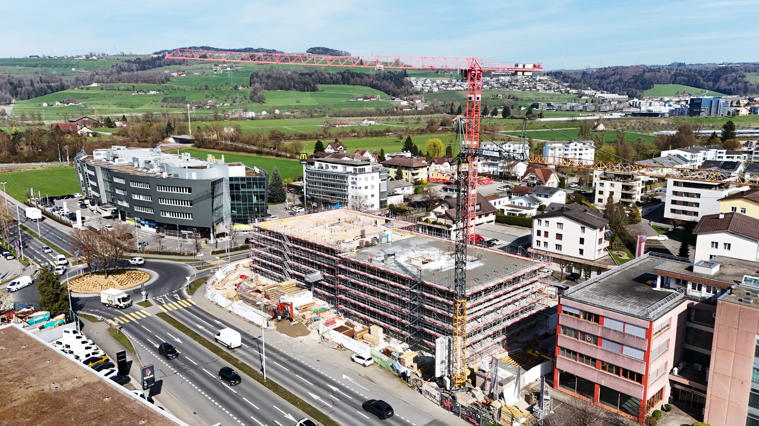 Aerial view of a construction site with a large red crane, surrounded by various buildings and a road with cars.