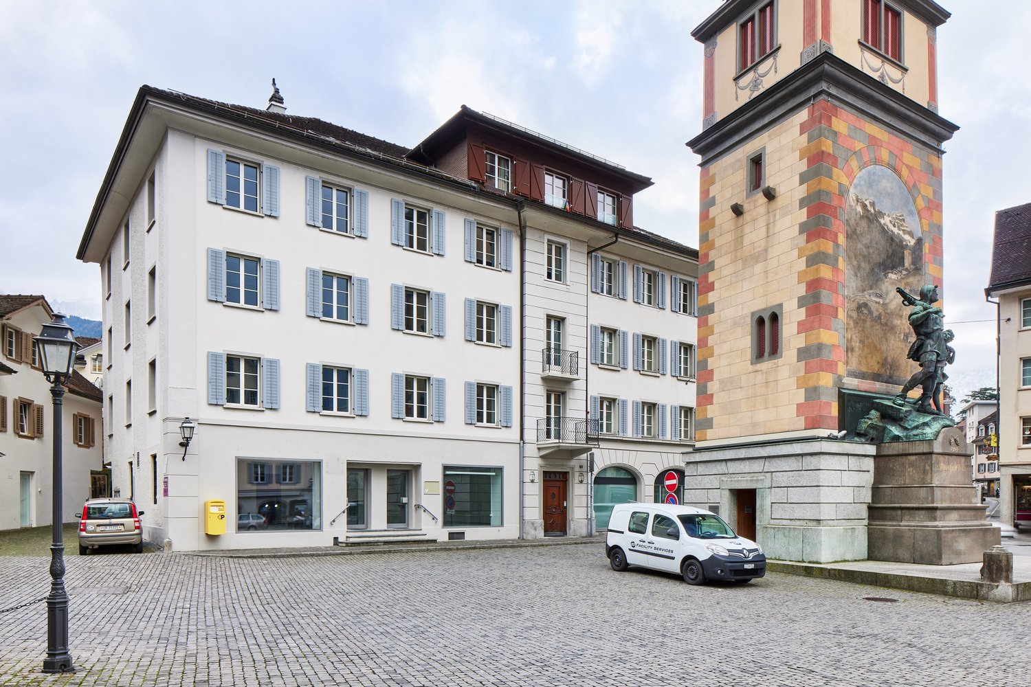 This image shows a historic town square with a tall clock tower and several multi-story buildings with traditional Swiss architectural styles. The buildings have white facades with blue shutters and balconies. There is a cobblestone plaza in the foregroun