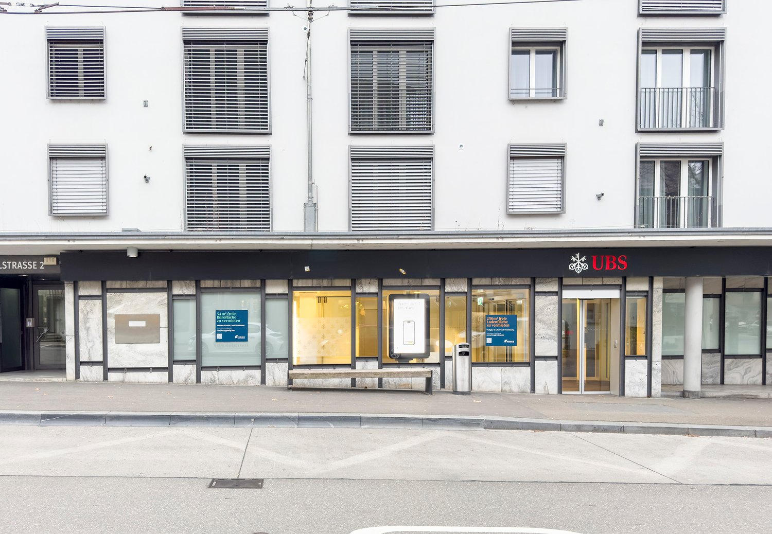 The image shows the exterior of a UBS bank branch. The building has a white facade with horizontal striped windows. The entrance is located in the center, with the UBS logo displayed prominently. There are several glass doors and windows, allowing visibil