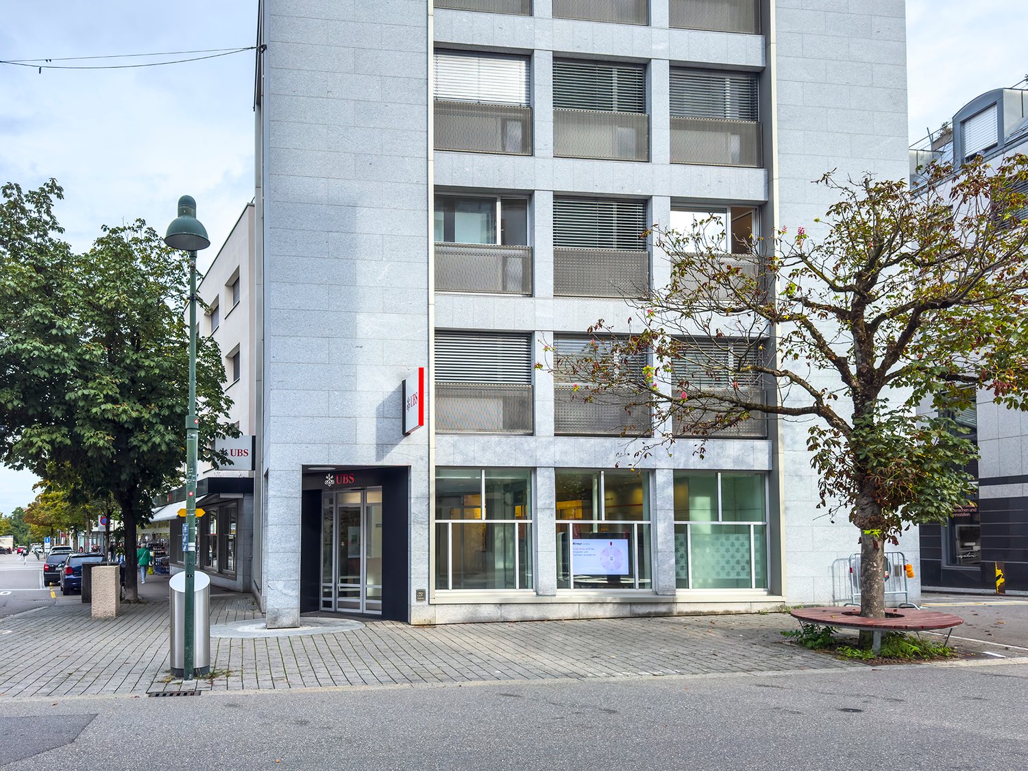 Multi-story commercial building with concrete exterior, large windows, and a UBS bank branch on the ground floor. The building is surrounded by trees and is located on a street with pedestrian walkways.