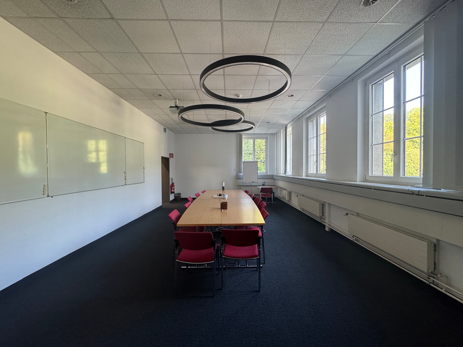Empty conference room with white walls, black carpet, glass whiteboards, and wooden long table. Large windows with white frames and red chairs.