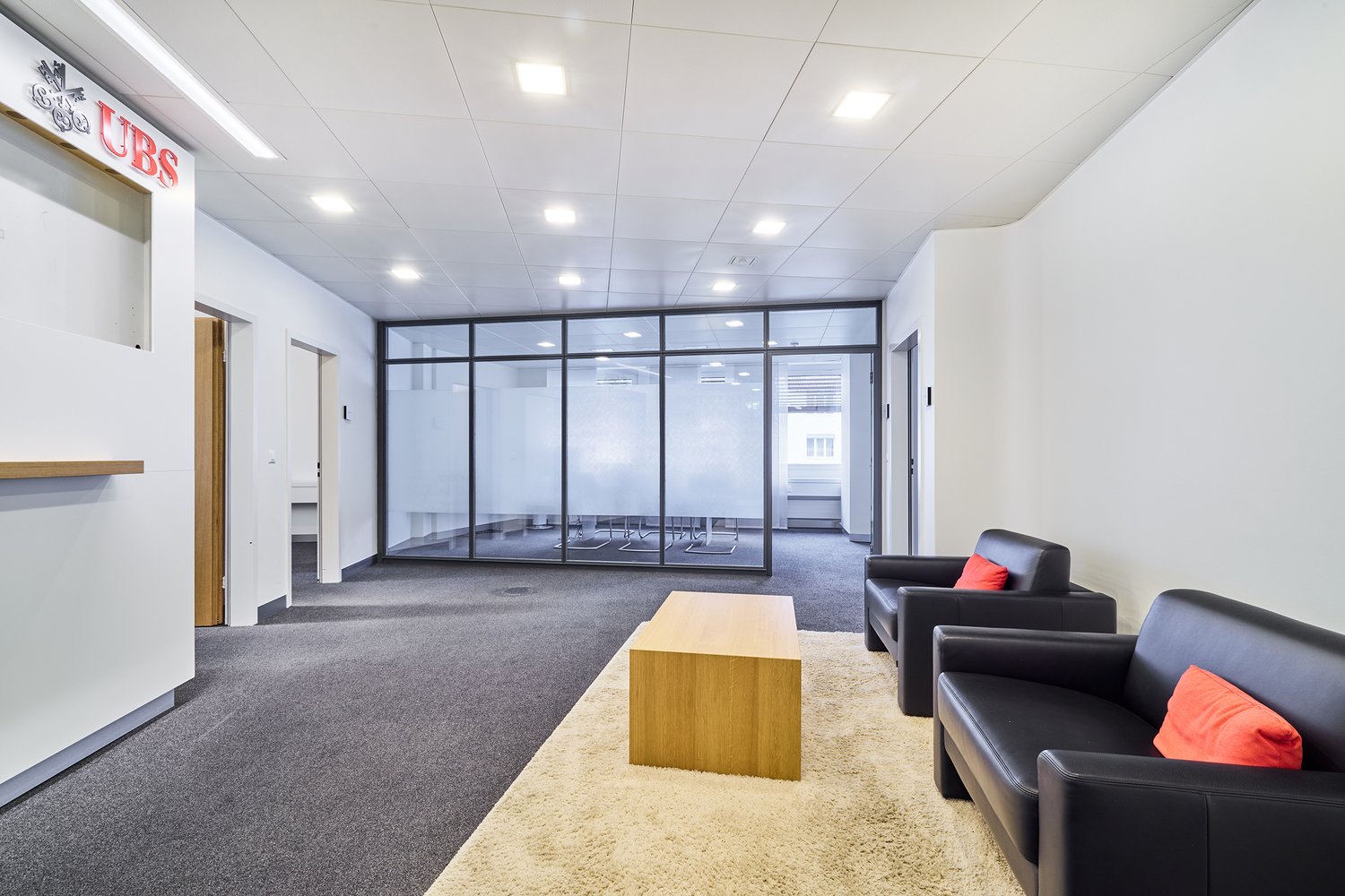 Carpeted floor, two black sofas with orange pillows, wooden coffee table, glass wall, open space, large white room