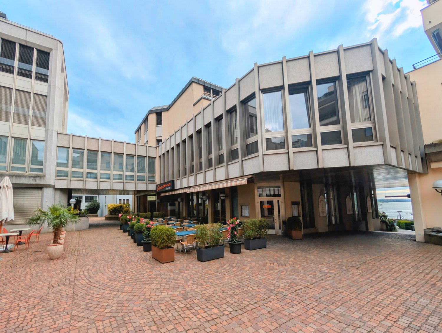 Modern building with glass windows, outdoor seating area, potted plants, brick pavement, and a view of the sea.
