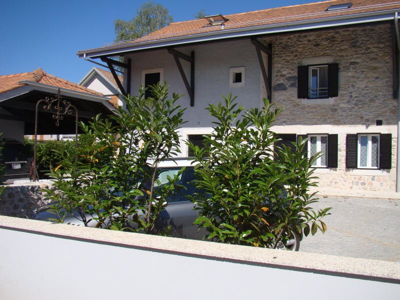 2-story stone and stucco house with red tile roof, large windows, and a balcony or terrace. The house is surrounded by lush greenery and plants.