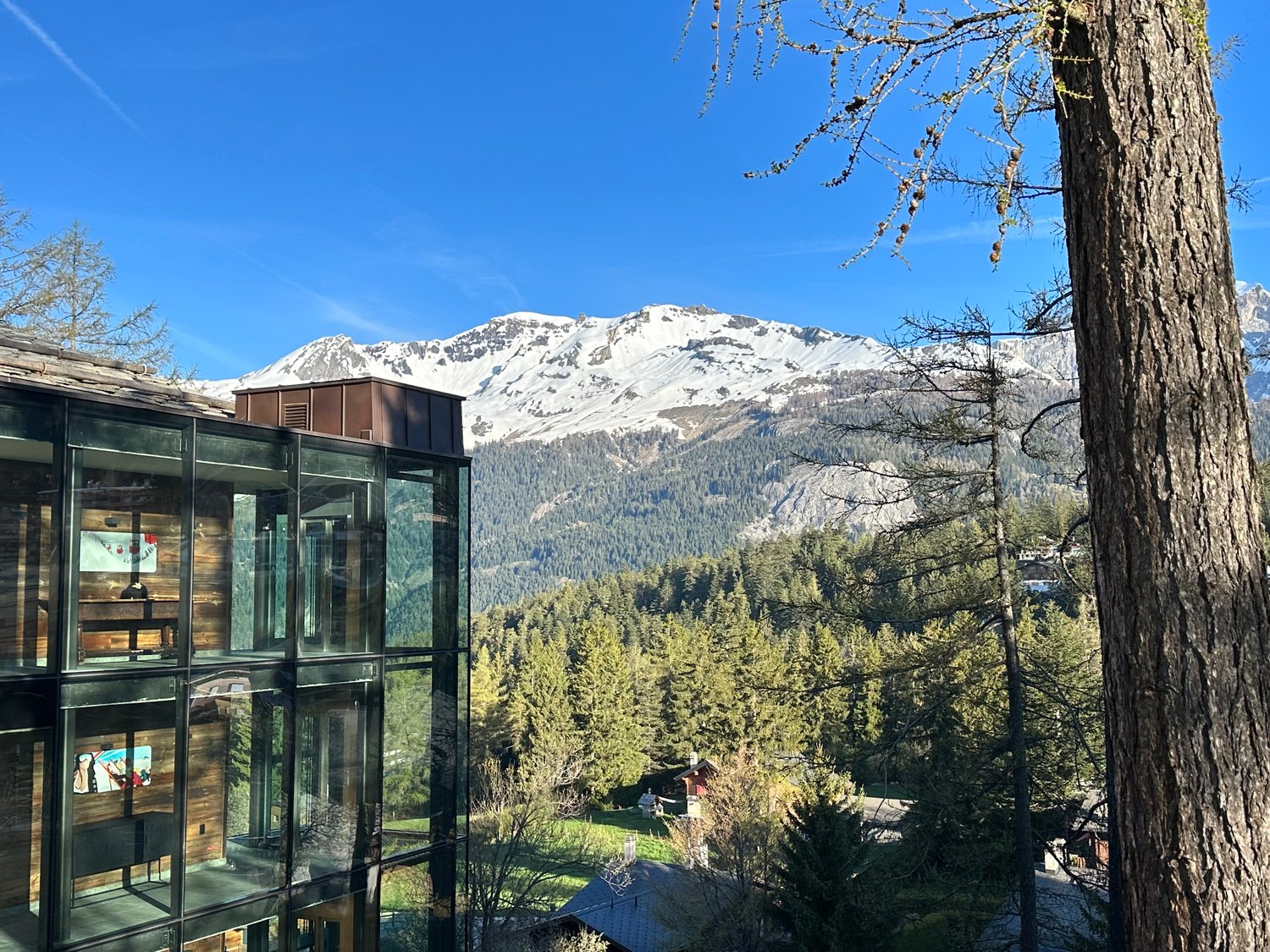 A glass-enclosed building with a view of snow-capped mountains and a forested valley in the background. The building appears to be situated in a scenic, mountainous area.