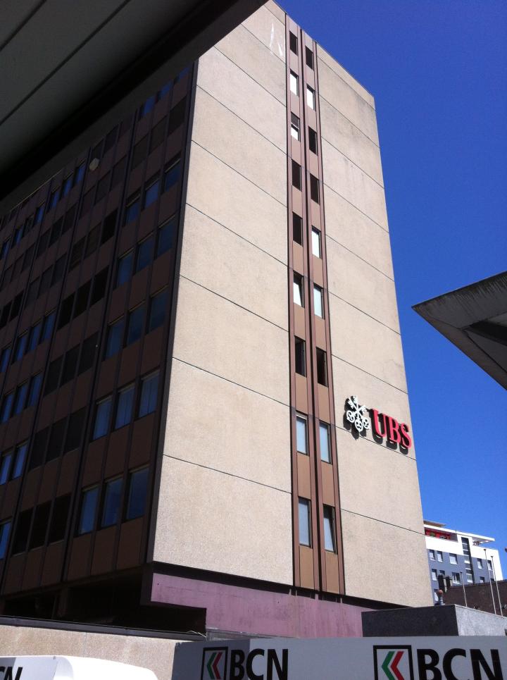 A multi-story commercial building with a prominent UBS logo on the facade. The building has a modern, angular design with large windows and a beige exterior. There is also a BCN logo visible on the building.