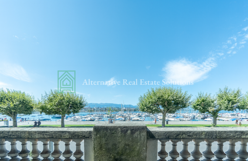 The image shows an outdoor area with a view of a marina or harbor. There are trees lining the area, and a stone balustrade or railing in the foreground. In the background, there are boats and yachts docked in the marina, with mountains visible in the dist