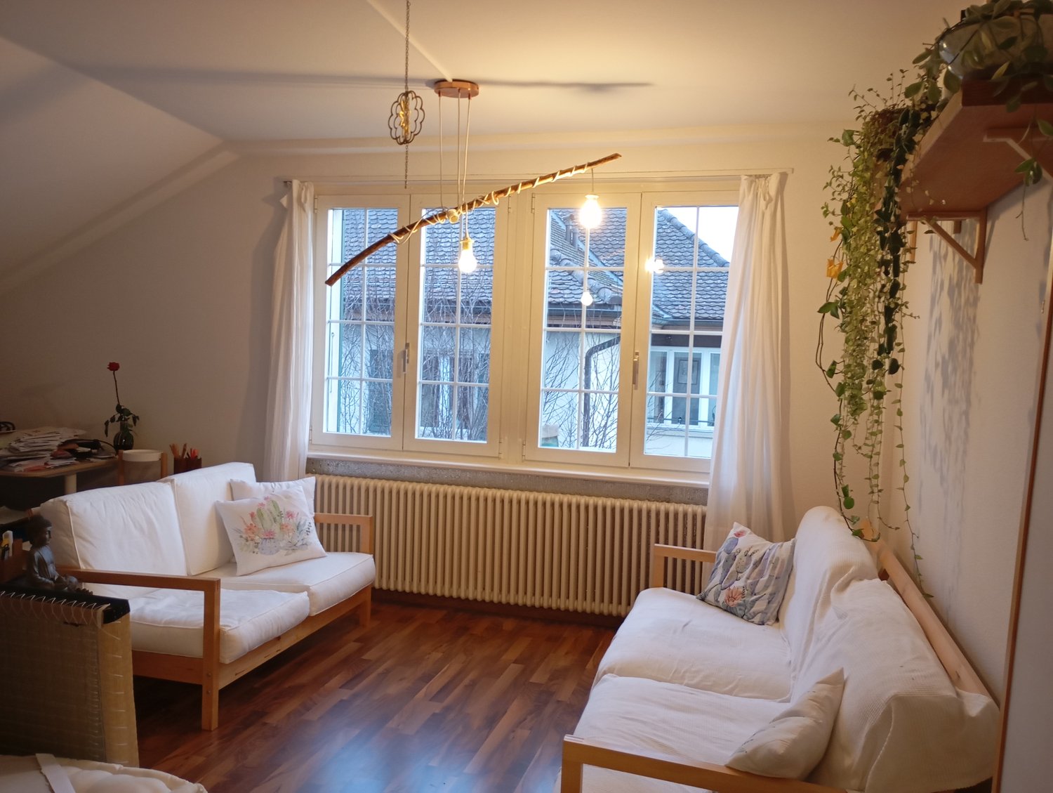 two white sofas, wooden coffee table, window with curtains, white radiator, wooden floor, ceiling with hanging light fixture, wall with hanging plant