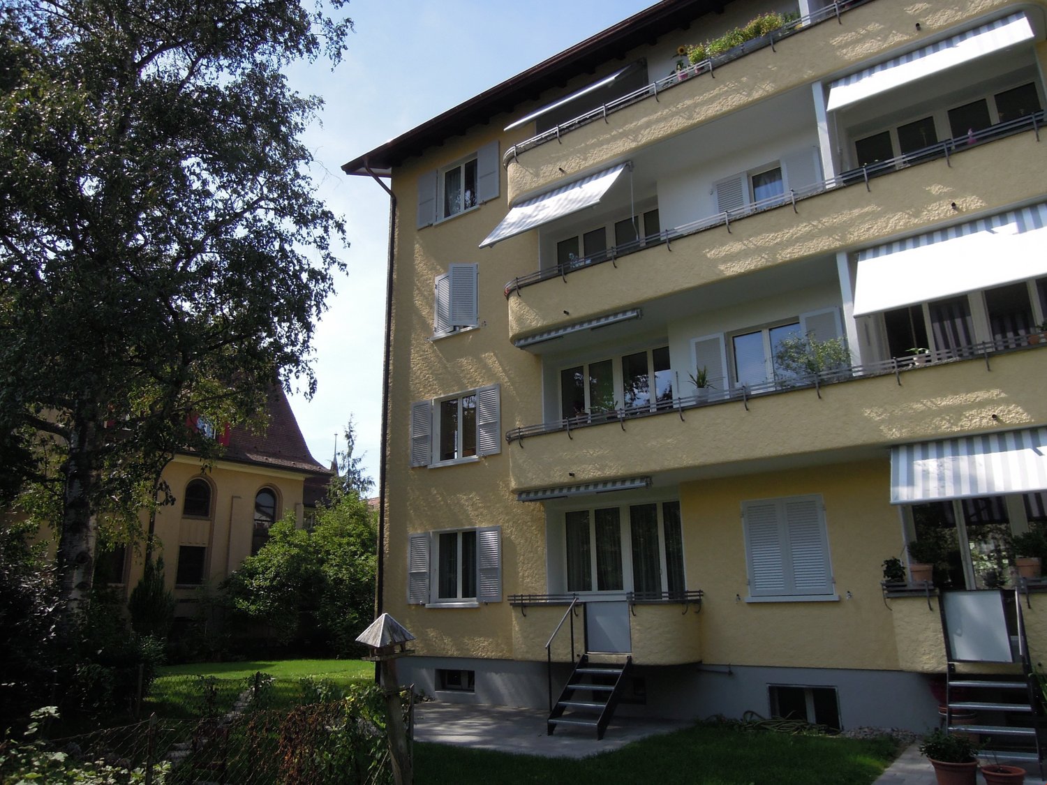 3-storey building with balconies, yellow paint, shrubs and trees in the surroundings, stairs leading to the entrance