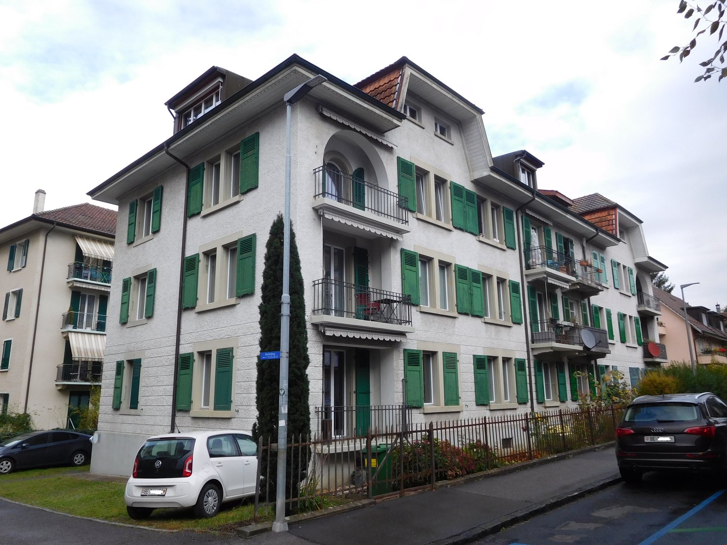 white apartment building with green shutters, balconies on the upper floors, two cars parked in front