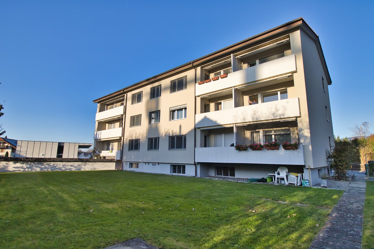 A multi-storey apartment building with beige exterior, large balcony with potted plants, front garden, and a view of trees in the distance.