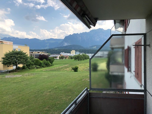 Glass door balcony, green grass, mountains, sky, building, tree, grass, and bushes.