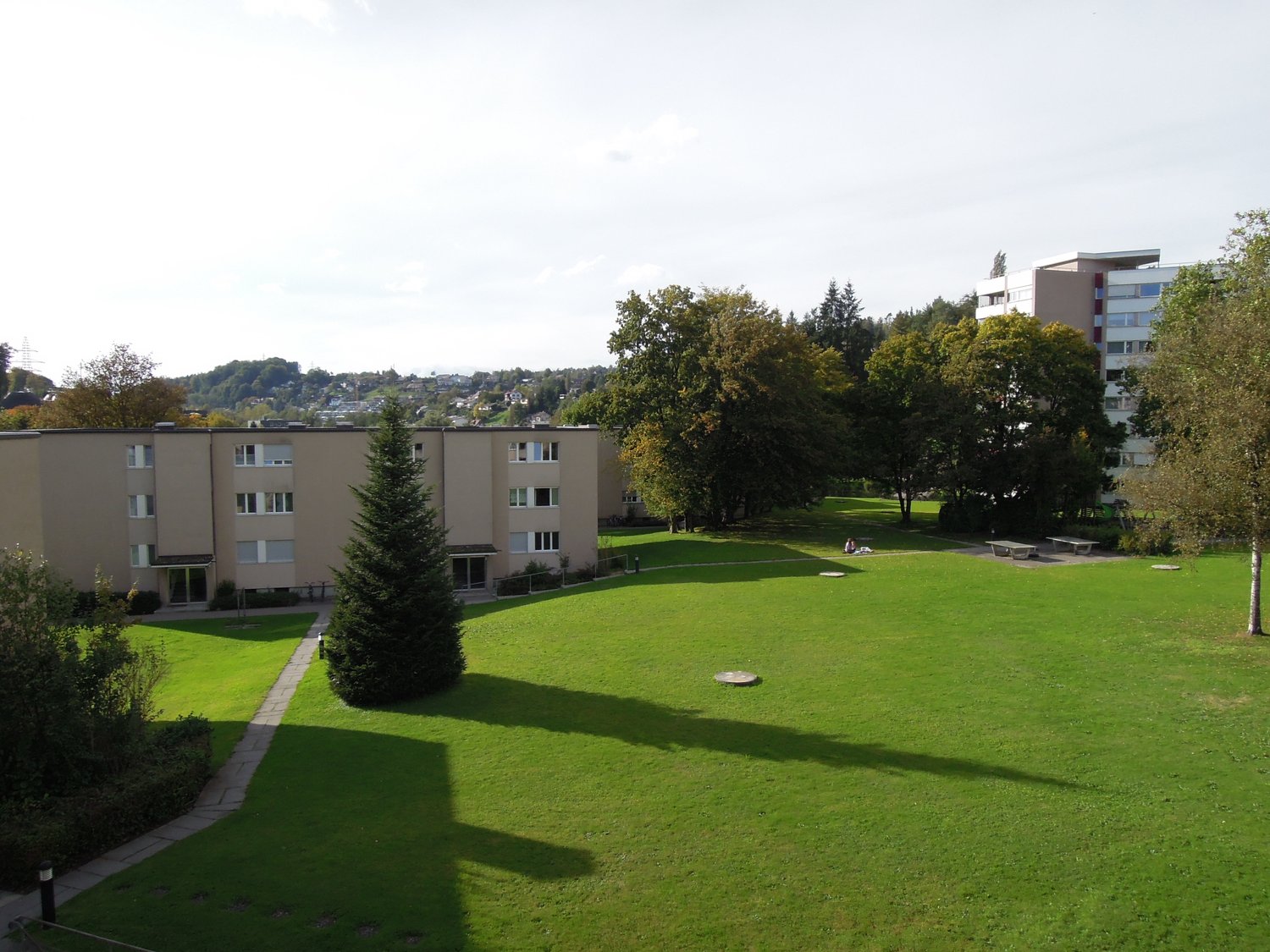 A three-story building with beige walls, multiple windows, a small pathway leading to a large lawn area with several trees and benches, and a distant view of a larger building.