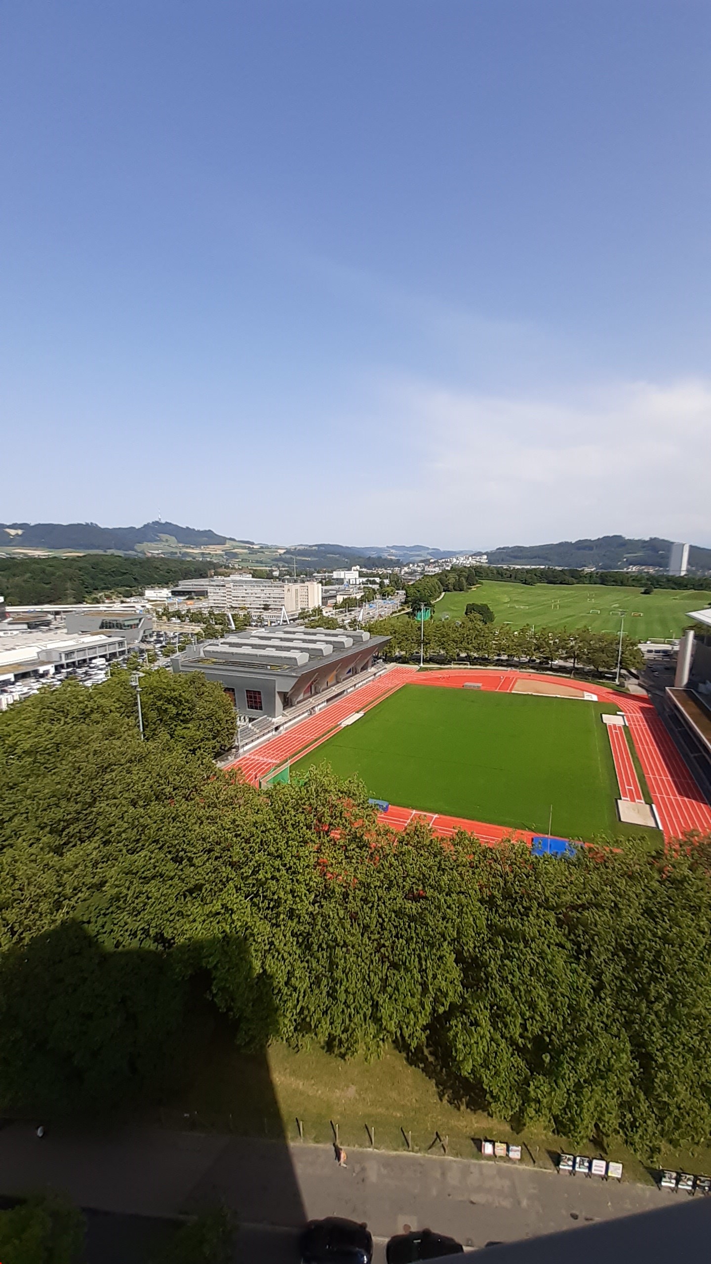 Aerial view of a wide green area, including a field, trees, and some buildings in the distance