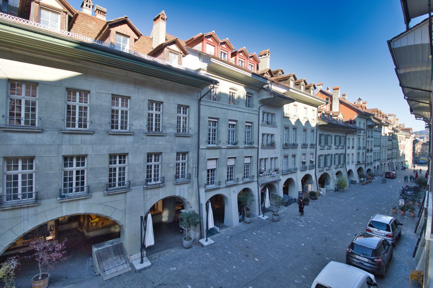 Cobblestone street, several parked cars, tall building with many windows, open archways with small shops, potted plants, people walking, trees