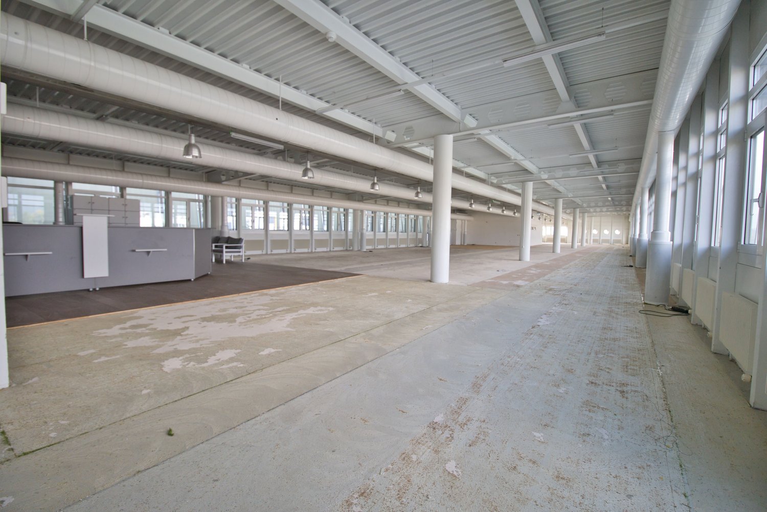 Empty building with high ceiling, large windows, concrete floor, metal beams, and pipes