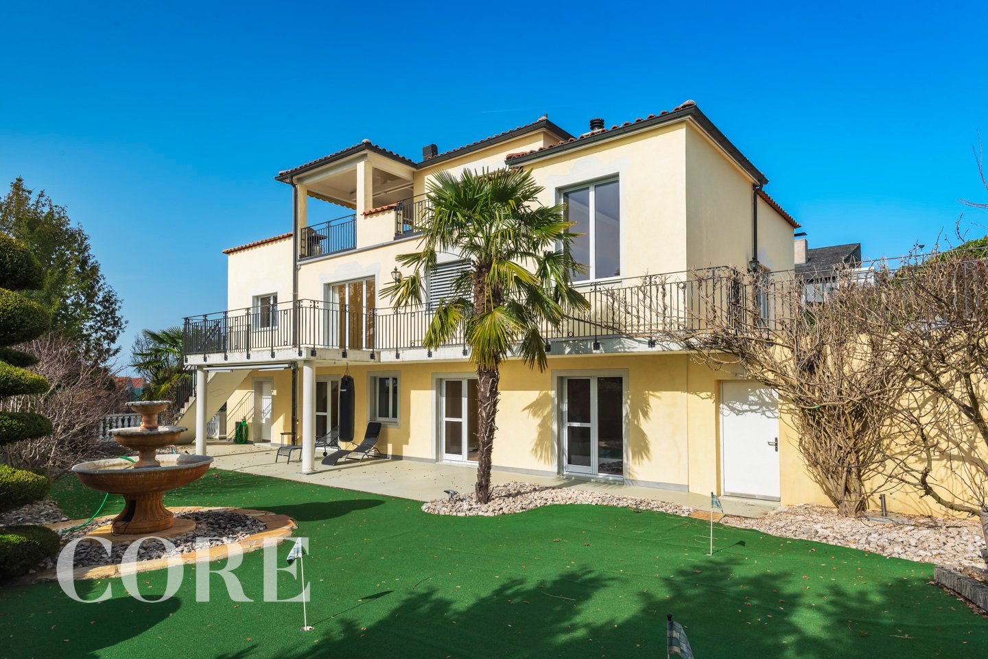 2 story house, yellow exterior, balcony, large windows, fountain, artificial grass lawn