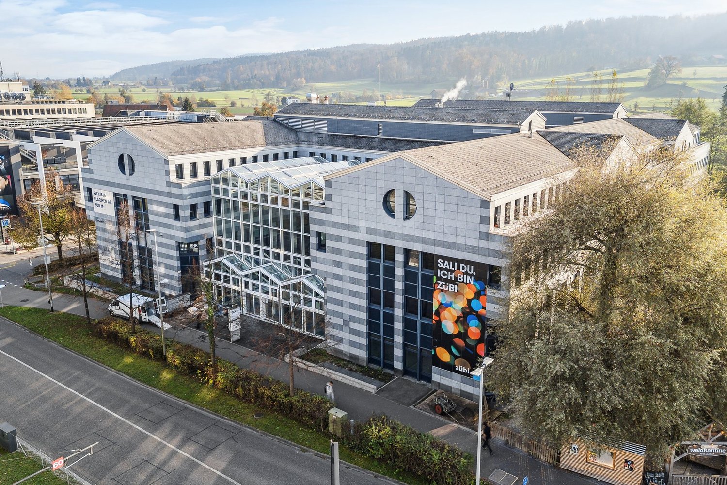 A large modern multi-story building with a glass facade and circular windows. The building has a sign that says 'Sali du ich bin zubi' on the side. The building is surrounded by trees and hills in the background, indicating a scenic location.