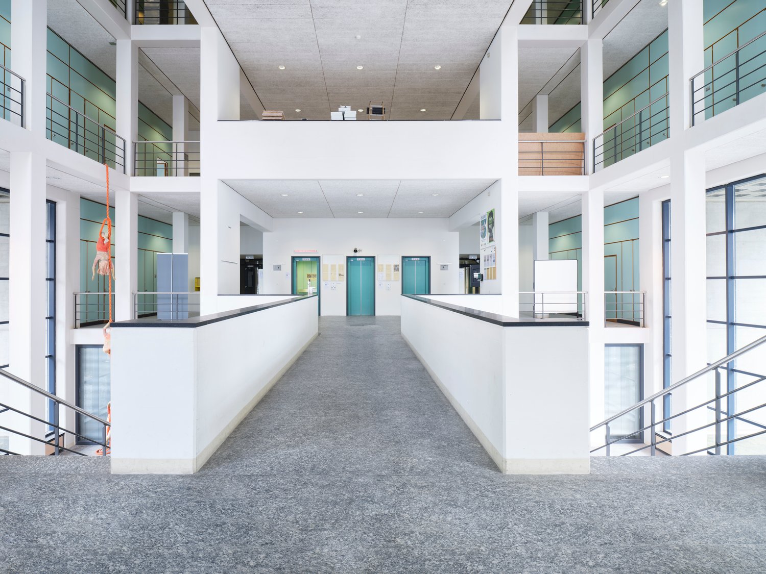 A modern office building interior with white walls, gray floors, glass windows, and metal railings on the balconies. Elevators are visible in the center, and there are two people in orange safety vests hanging from the ceiling.