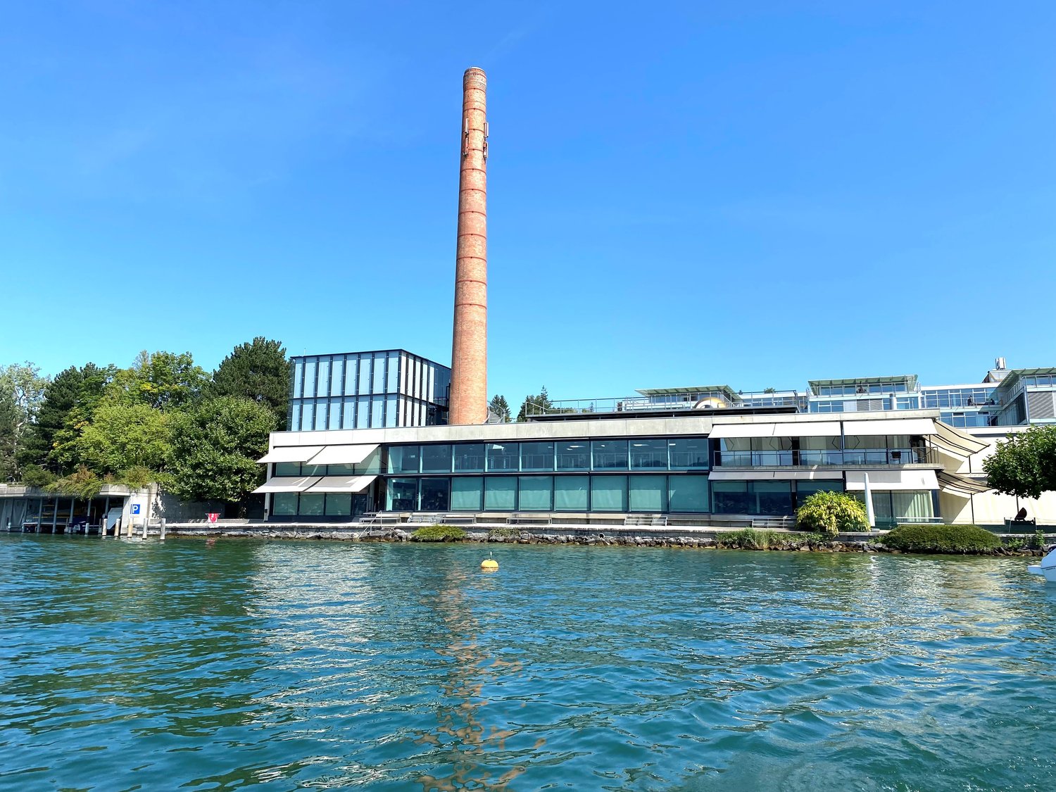 Large modern building with glass windows, overlooking water, tower in the center, surrounded by trees