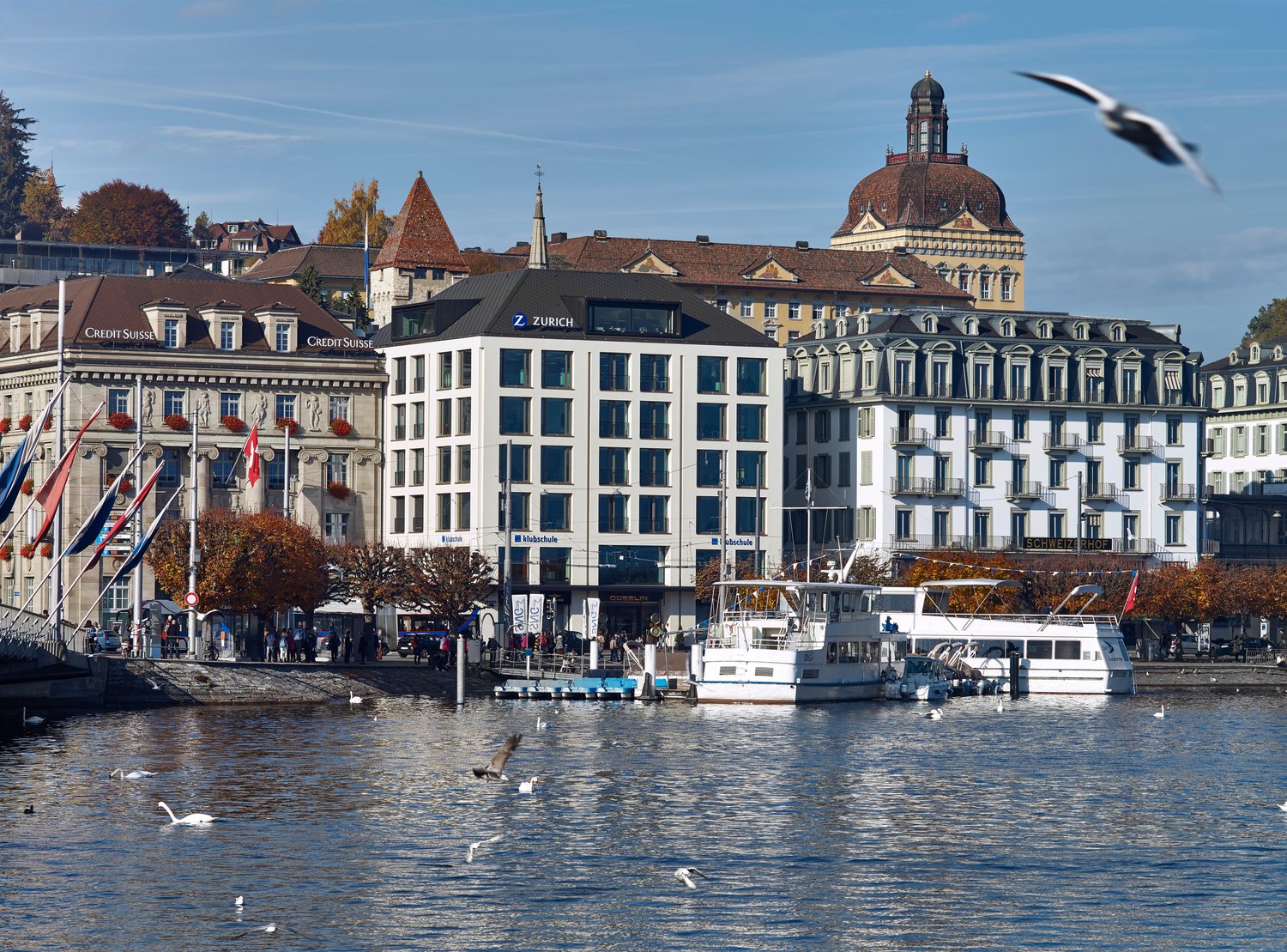 Urban waterfront with buildings, Credit Suisse logo, water, docked boats, people, flags
