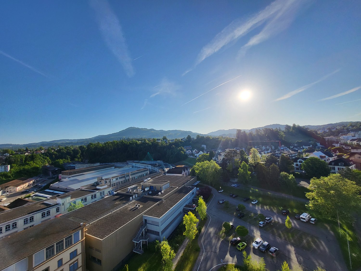 The image shows an aerial view of a town or city surrounded by mountains and forests. The town has a mix of residential and commercial buildings, with a river or stream running through the center. There are cars parked on the streets and trees lining the