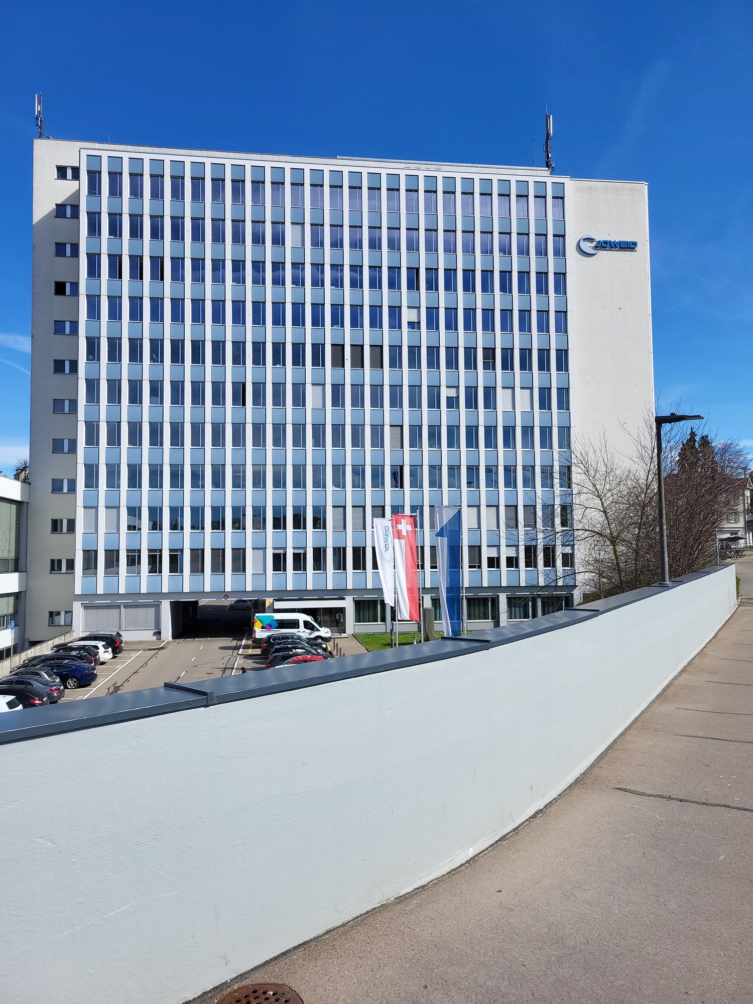 A large multi-story office building with a modern, rectangular design featuring numerous windows. The building has a prominent logo on the facade and is situated in an urban setting with a paved area in the foreground.