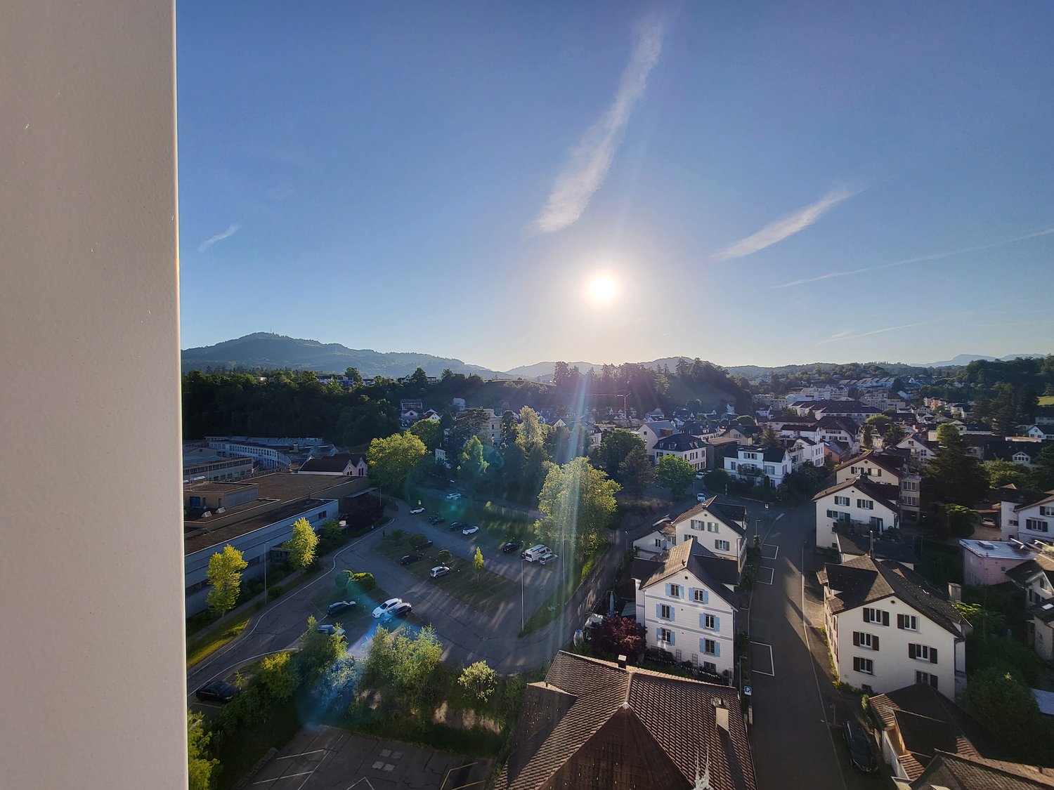 The image shows a panoramic view of a small town or city surrounded by hills and mountains. The town has a mix of residential and commercial buildings, with trees and greenery visible throughout. The sky is clear and blue, with the sun shining brightly. T