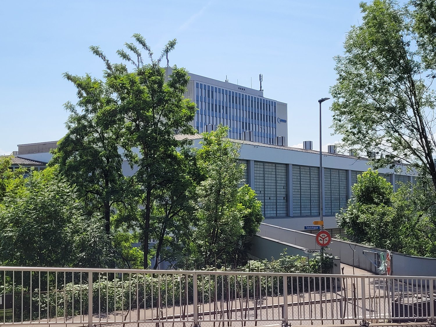 Large white building, several floors, glass windows, surrounded by trees, metal railings