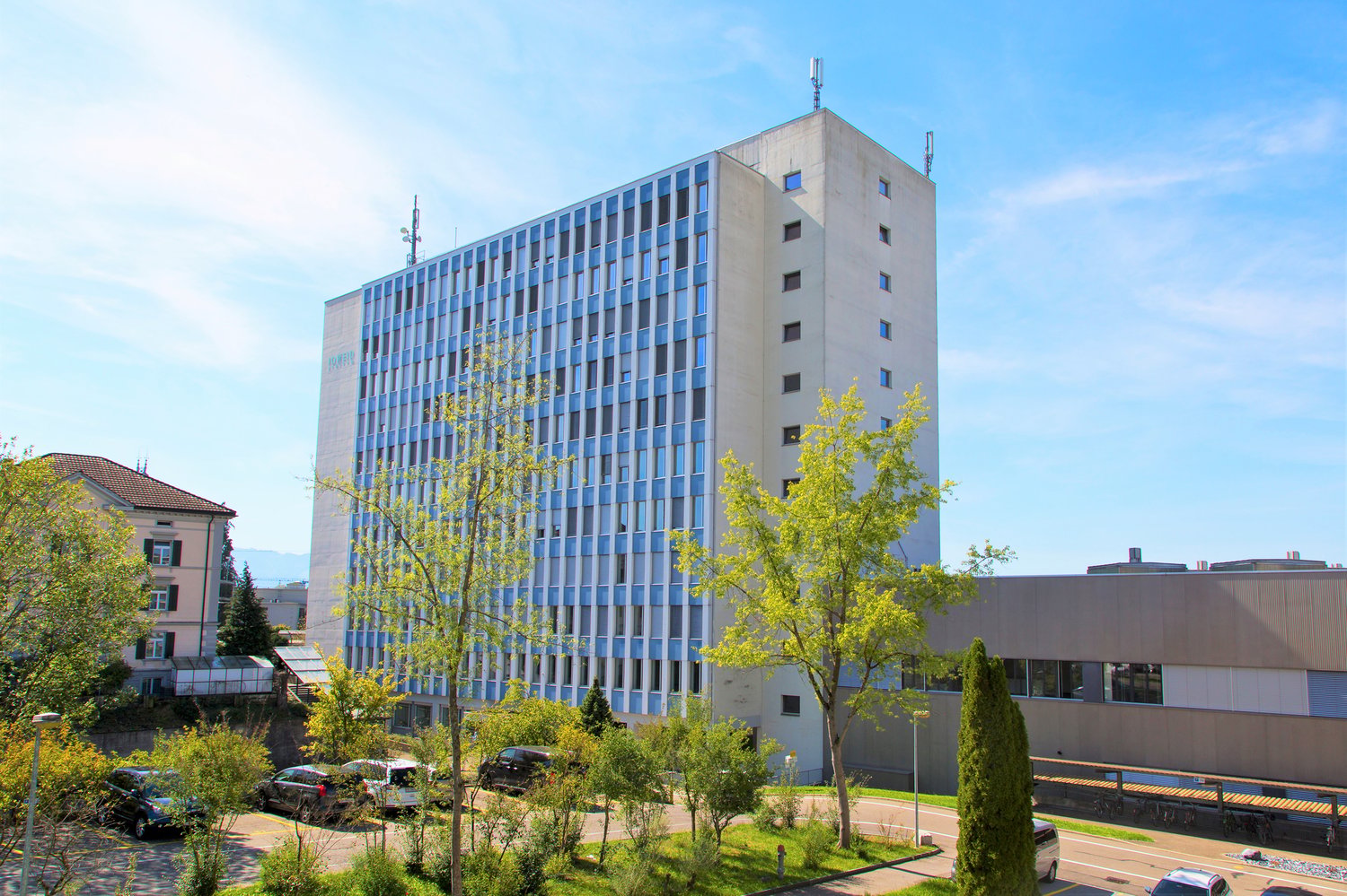 10 story white building, several windows, trees and bushes around, cars parked