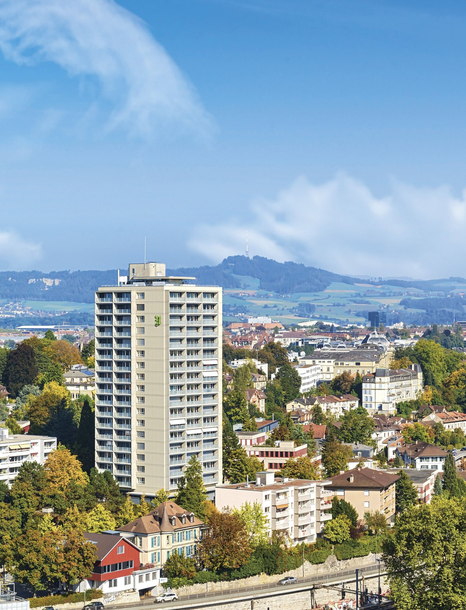 The image shows a cityscape with a tall apartment building in the foreground, surrounded by smaller residential buildings and trees. The city is situated in a hilly area with mountains visible in the background. The sky is blue with some clouds.