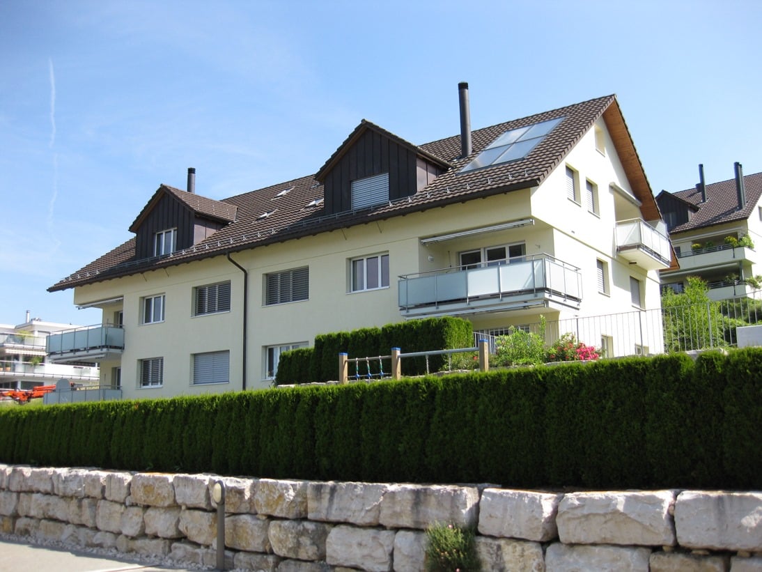 3-story apartment building with white exterior walls, black-tiled roof, and balconies on the upper floors. The building is surrounded by a stone wall and hedges.