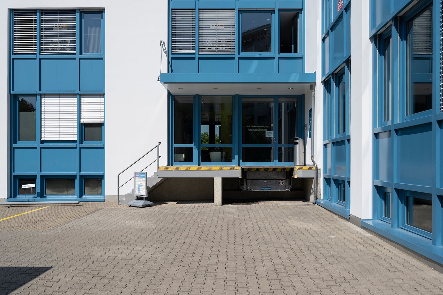 A modern commercial building with a raised ground floor and a parking area in front. The building has a blue exterior with large windows and a prominent entrance. The ground floor appears to be accessible via a ramp or platform, indicating a raised ground