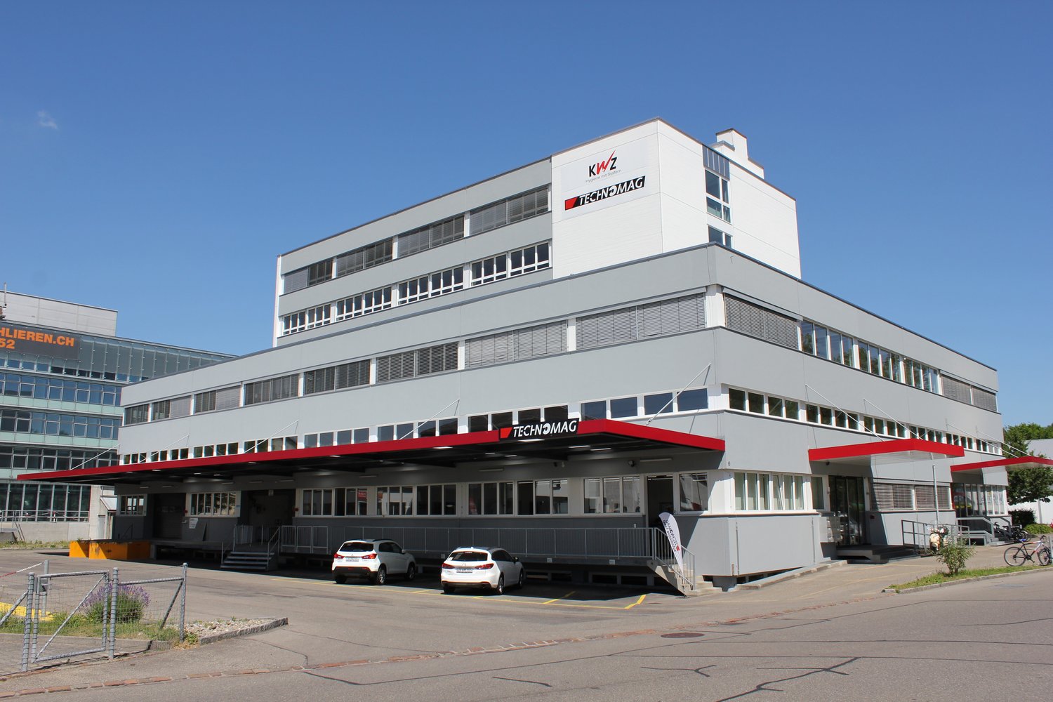 A multi-story commercial building with the Technomag logo prominently displayed. The building has large windows and a parking area in front.
