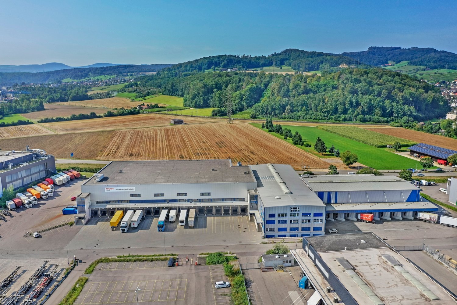 Large industrial building with multiple loading docks, surrounded by parking lots, trucks, and a vast green landscape.