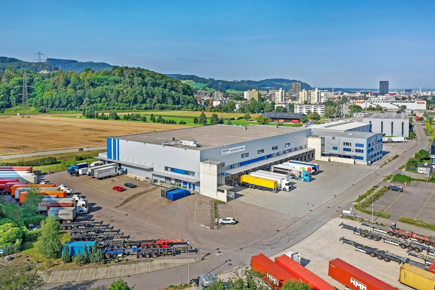 Large industrial building with multiple trucks parked in front, surrounded by a parking lot and a field.